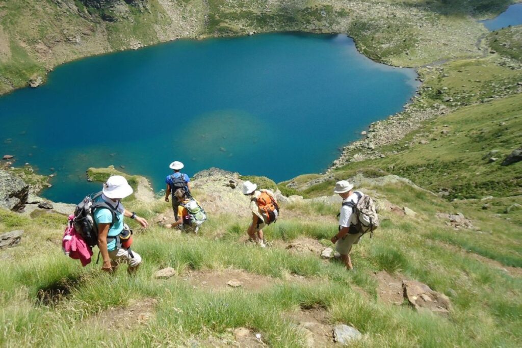 Groupe de marcheurs se dirigeant vers un lac de montagne en contrebas, le paysage est très verdoyant en raison de l'herbe en saison estivale et le lac ressort avec son bleu