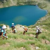 Groupe de marcheurs se dirigeant vers un lac de montagne en contrebas, le paysage est très verdoyant en raison de l'herbe en saison estivale et le lac ressort avec son bleu