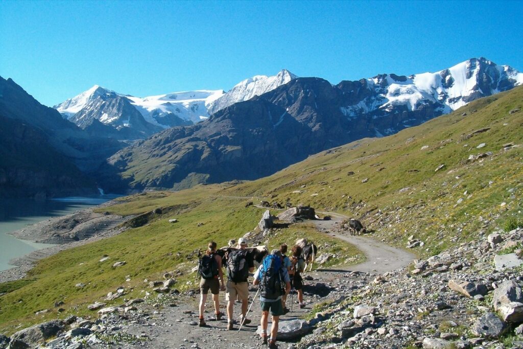 Griupe de randonneurs sur un entier de montagne avec face a eux des sommets couvert de neige et en contre bas sur la gauche un lac de montagne