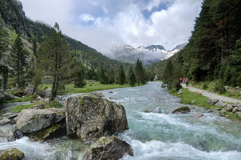 Paysage de montagne avec un cours d'eau en centre de l'image et des arbres des deux côtés, paysage qui s'ouvre sur la vallée et qui mène à des sommets