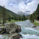 Paysage de montagne avec un cours d'eau en centre de l'image et des arbres des deux côtés, paysage qui s'ouvre sur la vallée et qui mène à des sommets