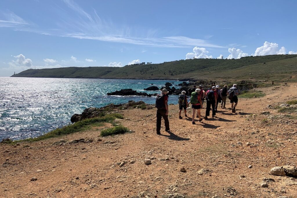 Groupe de randonneurs sur un sentier côtier de terre rouge avec à leur gauche la mer et face à eux les montagnes