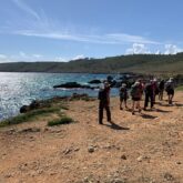 Groupe de randonneurs sur un sentier côtier de terre rouge avec à leur gauche la mer et face à eux les montagnes