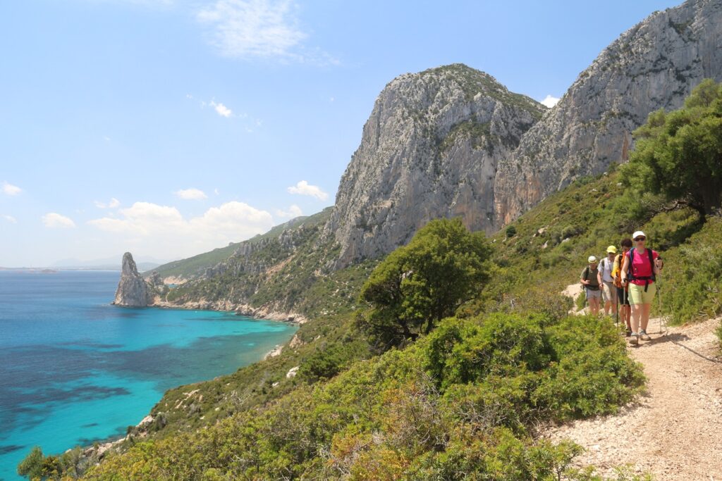 Groupe de randonneurs sur un sentier de randonnée en Sardaigne, parallèlement à ce sentier côtier, on voit la mer sur la gauche et des sommets en fond