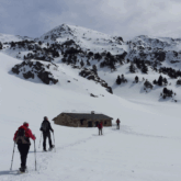 Groupe de marcheurs se dirigeant vers une cabane en pierre dans une étendue de neige avec un fond montagneux.