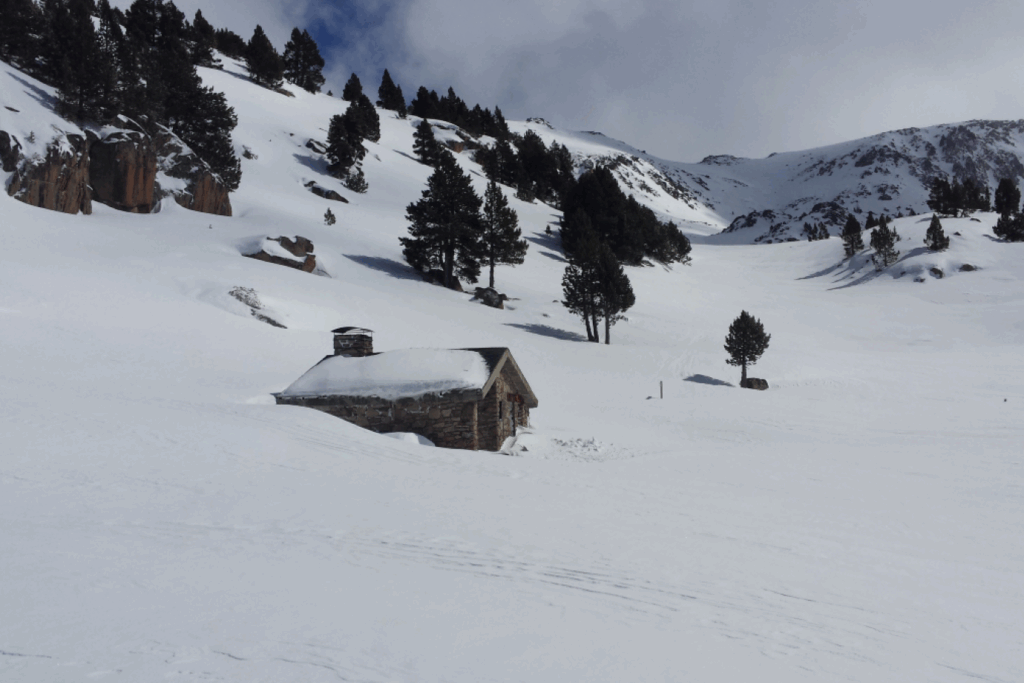 Paysage enneigé d'un flanc de montagne avec des arbres et une maison en pierre recouverte de neige, et des traces de randonneurs en raquettes.