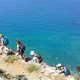 Groupe de randonneurs pris de haut sur un sentier côtier avec à leur droite la mer, l'eau a des couleurs turquoises, cette scène se déroule en Crète