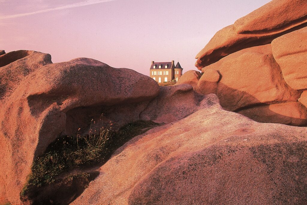Paysage sur la côte de granit rose en Bretagne avec un bâtiment en fond derrière les rochers du premier plan.