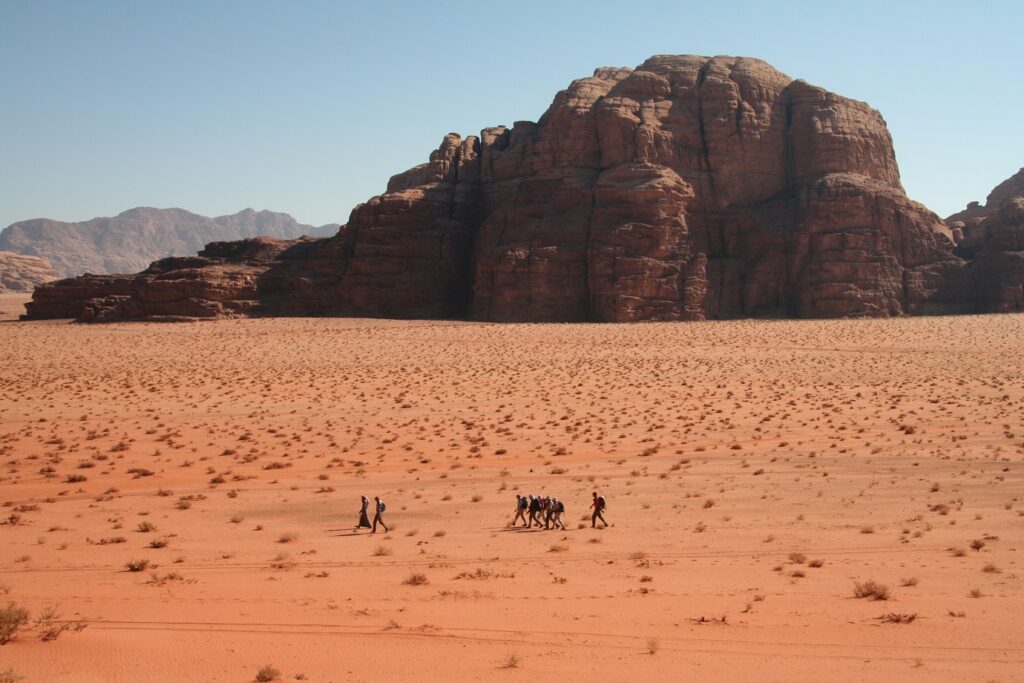 Groupe de randonneurs vu de loin qui marchent à travers le sable du désert du Wadi Rum avec en fond un sommet en pierre. On voit sur ce paysage de Jordanie beaucoup de couleurs orangées