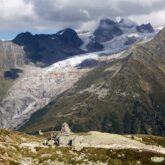 Paysage de montagne avec au premier plan un cairn de pierre et des anciennes constructions, et au-dessus des sommets et notamment le glacier