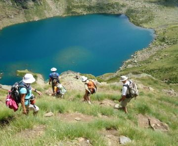 Groupe de marcheurs se dirigeant vers un lac de montagne en contrebas, le paysage est très verdoyant en raison de l'herbe en saison estivale et le lac ressort avec son bleu