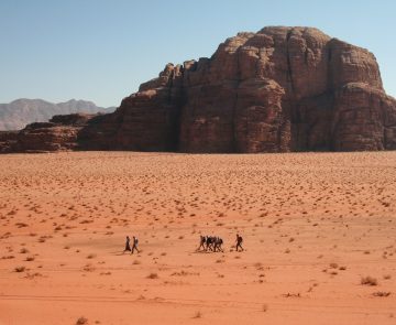 Groupe de randonneurs vu de loin qui marchent à travers le sable du désert du Wadi Rum avec en fond un sommet en pierre. On voit sur ce paysage de Jordanie beaucoup de couleurs orangées