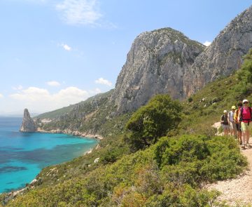 Groupe de randonneurs sur un sentier de randonnée en Sardaigne, parallèlement à ce sentier côtier, on voit la mer sur la gauche et des sommets en fond