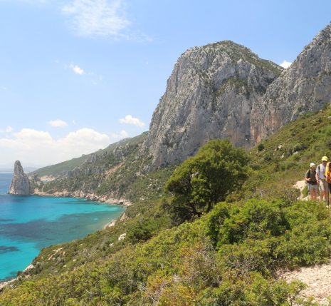 Groupe de randonneurs sur un sentier de randonnée en Sardaigne, parallèlement à ce sentier côtier, on voit la mer sur la gauche et des sommets en fond